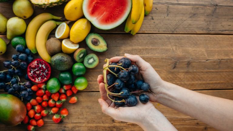 A variety of fresh, organic fruits with hands holding grapes on a wooden table.